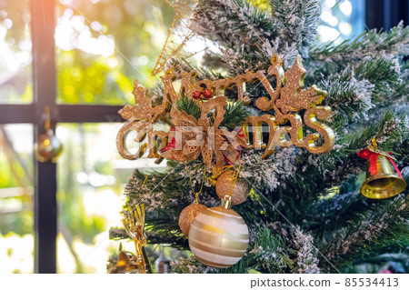 Golden shiny Merry Christmas handwriting logo on artificial pine tree for christmas time in coffee shop. Christmas tree decorations with color lights and merry christmas logo in cafe. Selective focus. 85534413