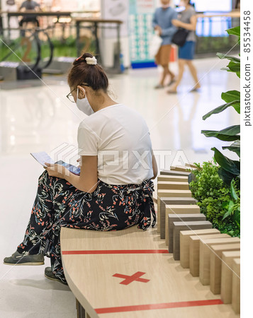 Back view of Asian woman wear face mask to prevent Corona virus (Covid-19) sit and using a smart phone and small notebook in public place and keep social distancing with other people. New normal. 85534458
