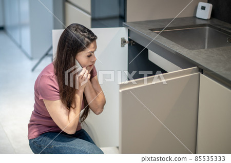 Confused woman with smartphone near ear looking under sink Confused woman with smartphone near ear looking under sink 85535333
