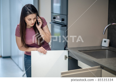 Woman looking under kitchen sink talking on smartphone Woman looking under kitchen sink talking on smartphone 85535334