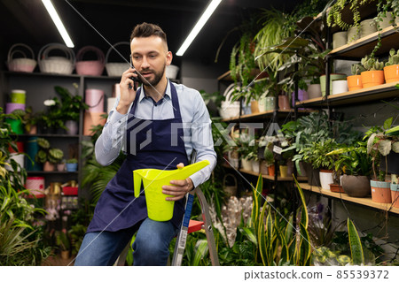 florist at work in a flower shop talking on the phone with a watering can in his hands florist at work in a flower shop talking on the phone with a watering can in his hands 85539372