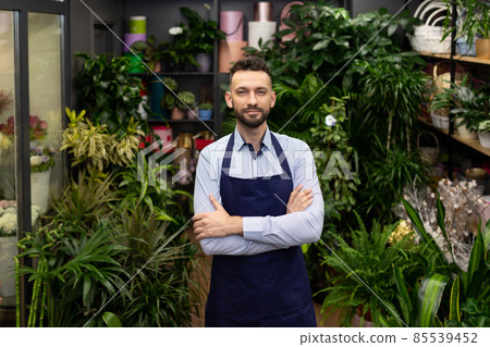 a florist man in a boutique of bouquets and flowers stands with a smile on his face in a blue apron 85539452