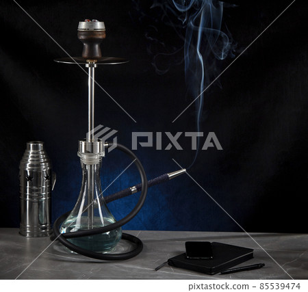 Beautiful modern hookah, from the mouthpiece of which smoke rises, stands on a table surrounded by a metal protective cap and a diary, on a dark background. Copy space. Beautiful modern hookah, from the mouthpiece of which smoke rises, stands on a table surrounded by a metal protective cap and a diary, on a dark background. Copy space. 85539474
