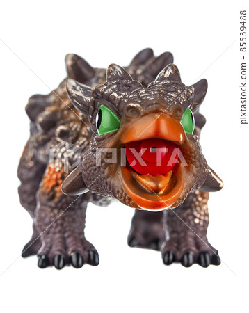Toy dinosaur of brownish color with green eyes, an open mouth and growths on the entire surface of the body, isolated on a white background. Toy dinosaur of brownish color with green eyes, an open mouth and growths on the entire surface of the body, isolated on a white background. 85539488