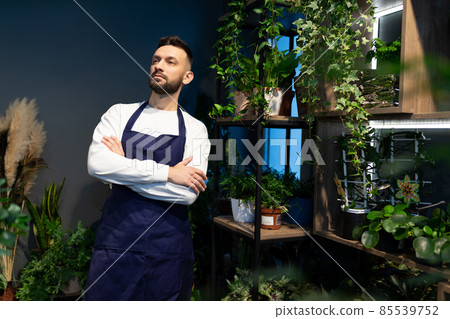 florist gardener in plant nurseries stands in a blue apron with a smile on his face florist gardener in plant nurseries stands in a blue apron with a smile on his face 85539752
