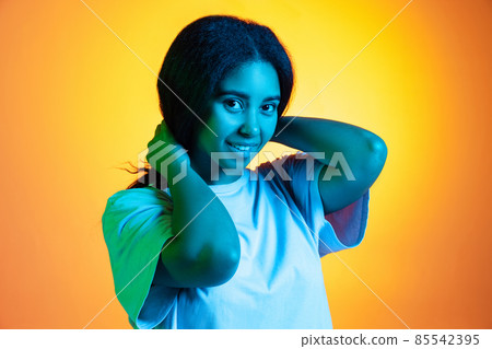 Excited young pretty dark skinned girl wearing white t-shirt isolated on yellow background in neon light, filter. 85542395