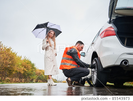 Roadside assistance worker replacing flat tire while beautiful woman in trench coat holding umbrella. Male auto mechanic changing flat tire on woman car the road. Concept of emergency road service. Roadside assistance worker replacing flat tire while beautiful woman in trench coat holding umbrella. Male auto mechanic changing flat tire on woman car the road. Concept of emergency road service. 85543973