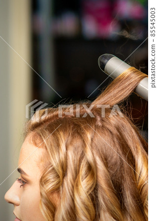 Hairdresser makes curls with a curling iron for the young woman with long brown hair in a beauty salon. 85545003