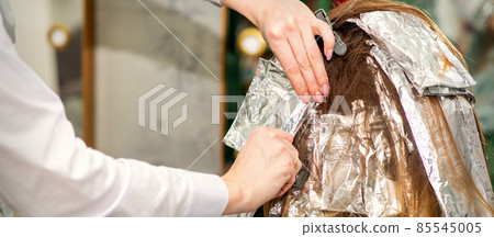 Hands of hairdresser dying hair applying foil for a brunette woman at the hairdressing salon, close up. 85545005