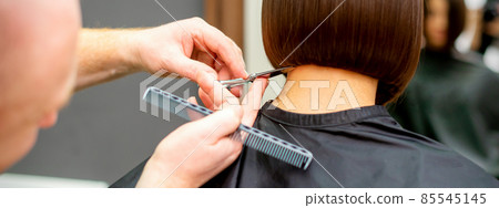 The hairdresser cuts the hair of a brunette woman. Hairstylist is cutting the hair of female client in a professional hair salon, close up. 85545145