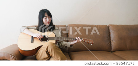 Asian woman in eyeglasses playing acoustic guitar while sitting on a couch at home. Hobbies and leisure activities concept. Copy space. 85547175