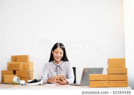 Young Asian woman business owner with many parcel boxes on the table happy online sales job, use your smartphone, get an order from customers, take notes, and make arrangements for delivery by post 85564399