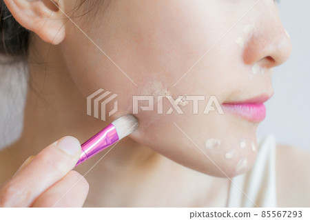 Closeup of young woman applying concealer on her facial skin with brush. Concealer is a type of cosmetic that is used to mask dark circles, age spots, large pores etc. Closeup of young woman applying concealer on her facial skin with brush. Concealer is a type of cosmetic that is used to mask dark circles, age spots, large pores etc. 85567293