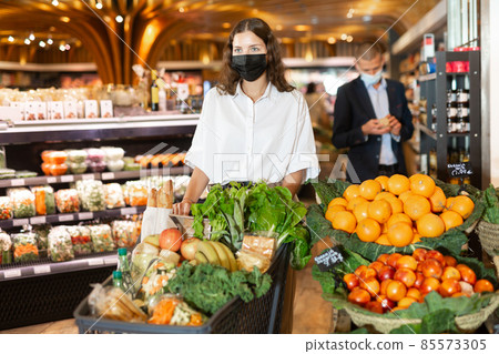 Portrait of a young girl in a protective mask with a grocery cart in the supermarket Portrait of a young girl in a protective mask with a grocery cart in the supermarket 85573305