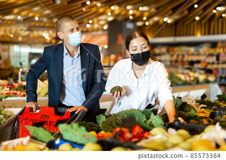Young couple wearing protective masks in the supermarket chooses avocados 85573384