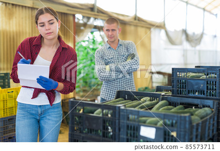 Female farmer working with papers in warehouse, male worker carrying crates with cucumber Female farmer working with papers in warehouse, male worker carrying crates with cucumber 85573711