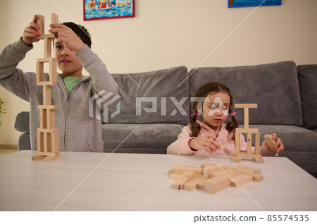 Adorable concentrated Caucasian kids sitting at table and playing board game, building tower with wooden blocks. Fine skill motors development, educational games, leisures, family relationship concept 85574535