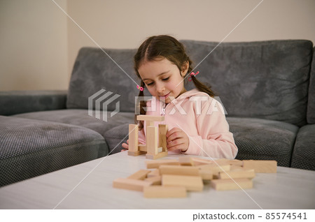 Cheerful creative beautiful Caucasian baby girl with two pigtails in pink sweatshirt playing educational developmental board game, she's concentrated on building wooden structures with blocks 85574541