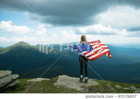 Female is waving American flag on top of mountain at sky background, celebrating independence day 85576607