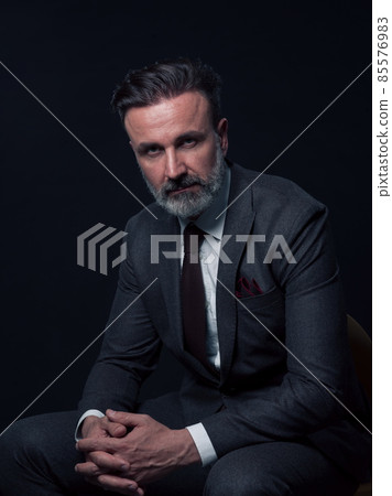 Portrait of adult businessman wearing trendy suit and sitting in modern studio on stylish chair against the black background Portrait of adult businessman wearing trendy suit and sitting in modern studio on stylish chair against the black background 85576983