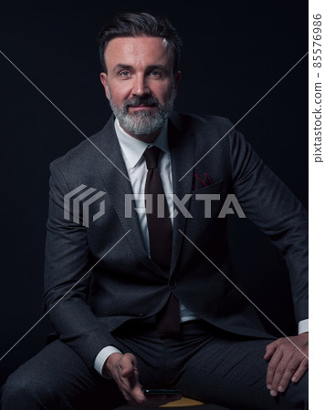Portrait of adult businessman wearing trendy suit and sitting in modern studio on stylish chair against the black background Portrait of adult businessman wearing trendy suit and sitting in modern studio on stylish chair against the black background 85576986
