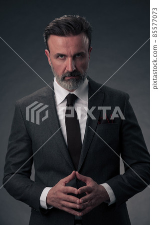 Portrait of a stylish elegant senior businessman with a beard and casual business clothes in photo studio isolated on dark background gesturing with hands Portrait of a stylish elegant senior businessman with a beard and casual business clothes in photo studio isolated on dark background gesturing with hands 85577073