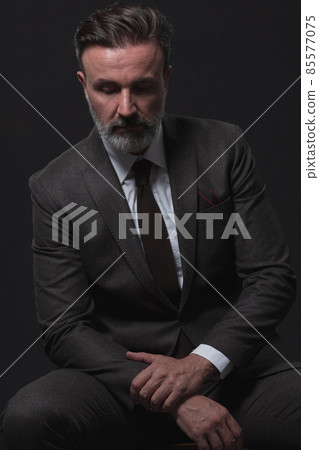 Portrait of adult businessman wearing trendy suit and sitting in modern studio on stylish chair against the black background Portrait of adult businessman wearing trendy suit and sitting in modern studio on stylish chair against the black background 85577075