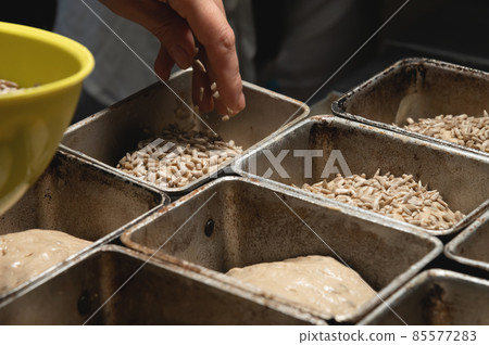 Close-up of female hands sprinkling a piece of raw dough in a baking form with seeds and sunflower. Prepare the dough before baking the bread. Craft bakery 85577283