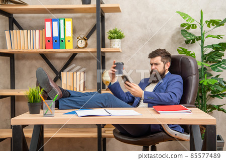 Serious man in suit relax in office chair reading sms in smartphone and drinking coffee, break Serious man in suit relax in office chair reading sms in smartphone and drinking coffee, break 85577489