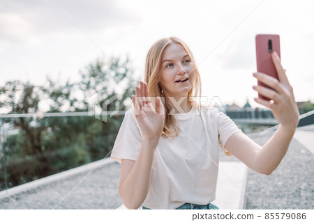 Blonde hair happy young woman posing outdoors in park take a selfie by mobile phone while standing in the park. Blonde hair happy young woman posing outdoors in park take a selfie by mobile phone while standing in the park. 85579086