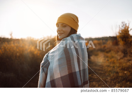 Outdoor woman with a blanket on her shoulders smiles enjoys sunset in autumn nature meadow at sunset in nature. Hope, mental strength concept. Outdoor woman with a blanket on her shoulders smiles enjoys sunset in autumn nature meadow at sunset in nature. Hope, mental strength concept. 85579087