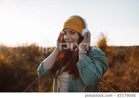 Stylish 30s caucasian female in headphones listening music, enjoys music in the meadow 85579088