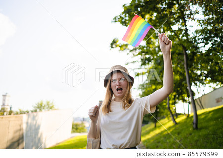Beautiful caucasian lesbian woman smiling and holding rainbow flag during pride parade walking on city street 85579399