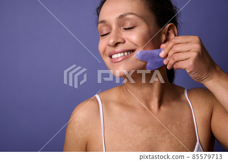 Beauty portrait of stunning delightful self-confident woman with skin problems and vitiligo smiling toothy smile massaging her face with Ayurvedic Gua-Sha jade massager against violet background Beauty portrait of stunning delightful self-confident woman with skin problems and vitiligo smiling toothy smile massaging her face with Ayurvedic Gua-Sha jade massager against violet background 85579713