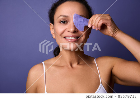 Beautiful half-naked African woman smiles toothy smile, holds Ayurvedic Gua Sha jade stone massager near her face, looking at the camera, posing on purple background with a copy space for advertising Beautiful half-naked African woman smiles toothy smile, holds Ayurvedic Gua Sha jade stone massager near her face, looking at the camera, posing on purple background with a copy space for advertising 85579715