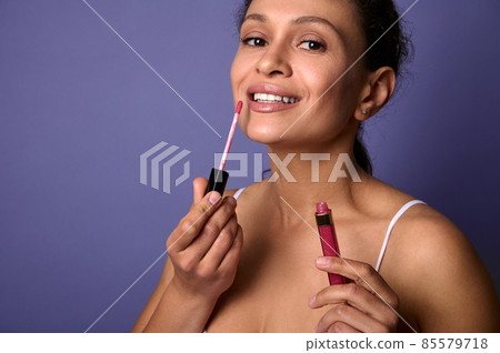 Attractive natural beauty African woman applies lipstick, hygienic lip gloss to plump lips, smiles toothy smile looking at camer, posing over purple background with copy space for ads. Make-up concept Attractive natural beauty African woman applies lipstick, hygienic lip gloss to plump lips, smiles toothy smile looking at camer, posing over purple background with copy space for ads. Make-up concept 85579718