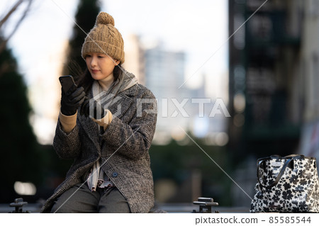 A woman sitting on an outdoor bench at dusk and operating a smartphone 85585544