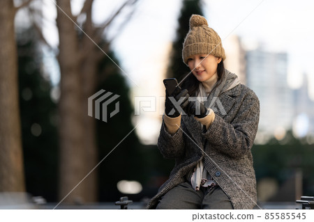 A woman sitting on an outdoor bench at dusk and operating a smartphone 85585545