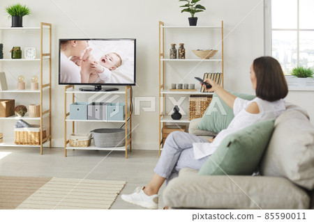 Woman sitting on sofa at home and watching movie on television. Happy relaxed adult lady resting on couch in living room interior and enjoying her favorite show on modern TV set with big screen Woman sitting on sofa at home and watching movie on television. Happy relaxed adult lady resting on couch in living room interior and enjoying her favorite show on modern TV set with big screen 85590011
