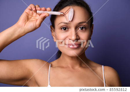 Beautiful middle aged woman with healthy perfect clean skin and natural makeup massaging her face with pink stone jade roller massager, smiles looking at camera, isolated over violet background 85592812