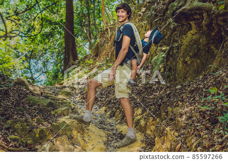 The father carries his son in a baby carrying is hiking in the forest. Tourist is carrying a child on his back in the nature of Vietnam The father carries his son in a baby carrying is hiking in the forest. Tourist is carrying a child on his back in the nature of Vietnam 85597266