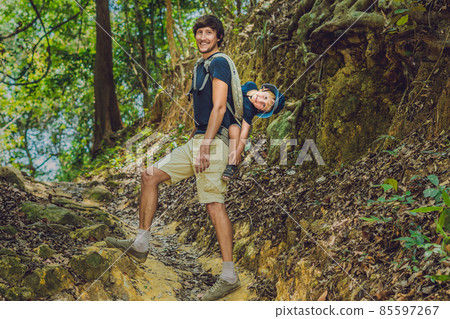 The father carries his son in a baby carrying is hiking in the forest. Tourist is carrying a child on his back in the nature of Vietnam The father carries his son in a baby carrying is hiking in the forest. Tourist is carrying a child on his back in the nature of Vietnam 85597267