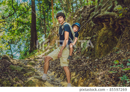 The father carries his son in a baby carrying is hiking in the forest. Tourist is carrying a child on his back in the nature of Vietnam The father carries his son in a baby carrying is hiking in the forest. Tourist is carrying a child on his back in the nature of Vietnam 85597269