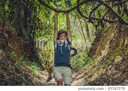 The father carries his son in a baby carrying is hiking in the forest. Tourist is carrying a child on his back in the nature of Vietnam The father carries his son in a baby carrying is hiking in the forest. Tourist is carrying a child on his back in the nature of Vietnam 85597270