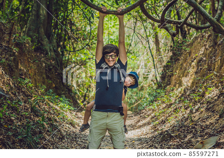 The father carries his son in a baby carrying is hiking in the forest. Tourist is carrying a child on his back in the nature of Vietnam The father carries his son in a baby carrying is hiking in the forest. Tourist is carrying a child on his back in the nature of Vietnam 85597271