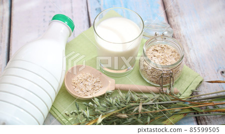 Close-up of a glass and a bottle with oat milk, oat spikelets, and a wooden spoon with oat flakes on a light table . Alternative vegan milk Close-up of a glass and a bottle with oat milk, oat spikelets, and a wooden spoon with oat flakes on a light table . Alternative vegan milk 85599505