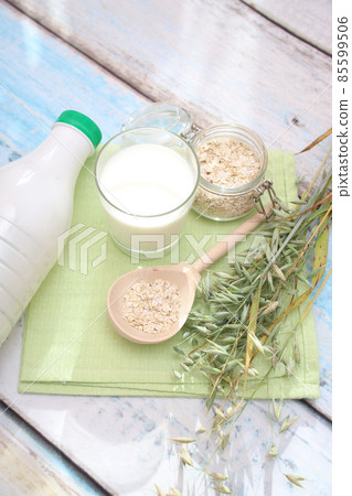 A glass of oat milk, oat spikelets, a wooden spoon with oat flakes and a bottle on a bright table . Alternative vegan milk 85599506