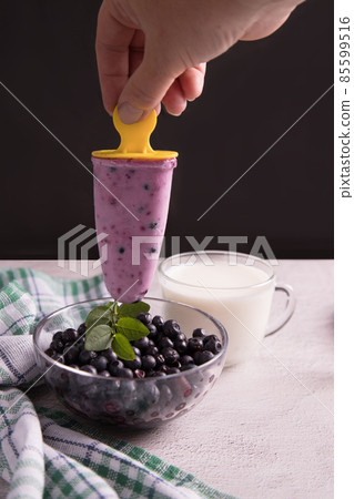 A woman's hand holds homemade ice cream with berries over a plate of blueberries on a dark background. The concept of cooking homemade food 85599516