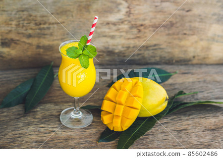 Juicy smoothie from mango in glass with striped red straw and with a mint leaf on old wooden background. Healthy life concept, copy space Juicy smoothie from mango in glass with striped red straw and with a mint leaf on old wooden background. Healthy life concept, copy space 85602486
