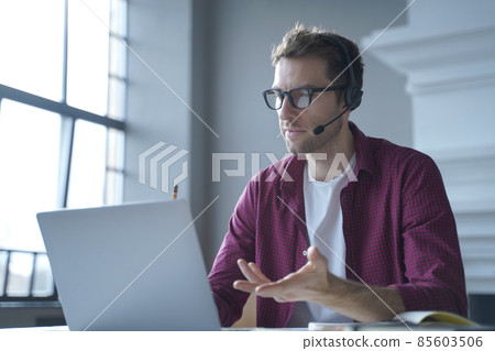 European man online tutor wearing glasses sitting at desk in headset and talking by video call 85603506